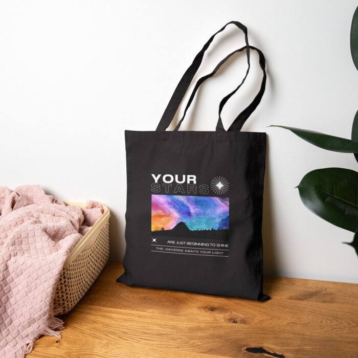 Black canvas tote bag featuring a vibrant cosmic sky design, displayed on a wooden surface next to a basket.