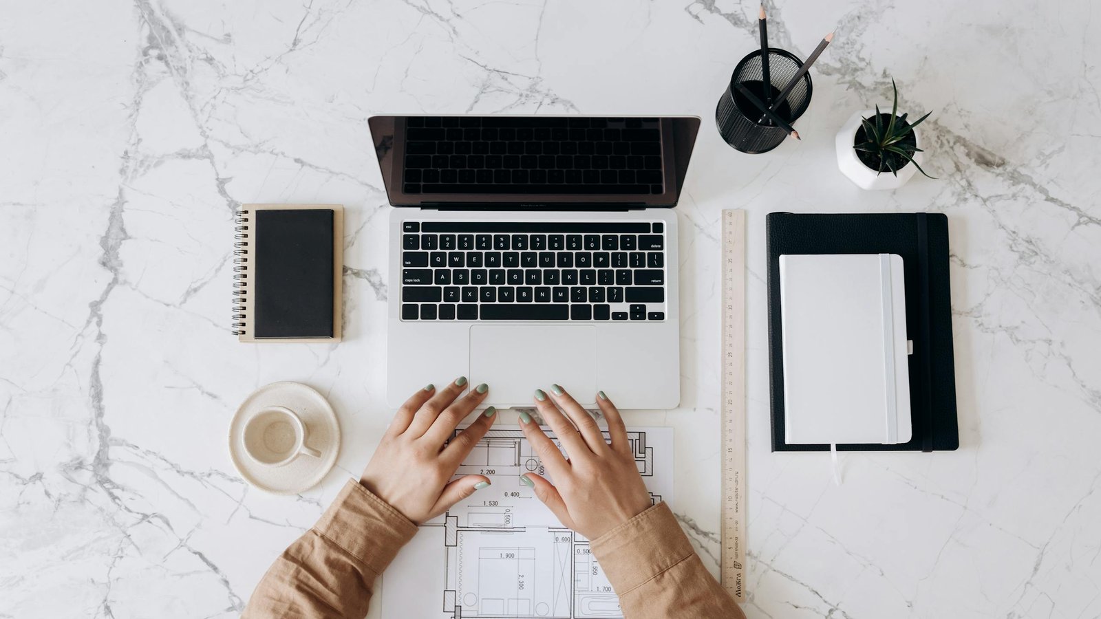 Photo by Mikhail Nilov | WISE360 Top view of a stylish home office desk with a laptop, planner, and coffee cup, showing hands on a blueprint.